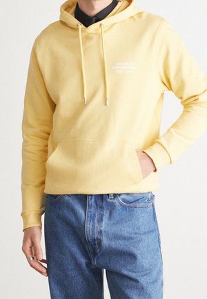 Man wearing yellow hoodie with front pocket, blue jeans, and black shirt underneath, standing with one hand in pocket.