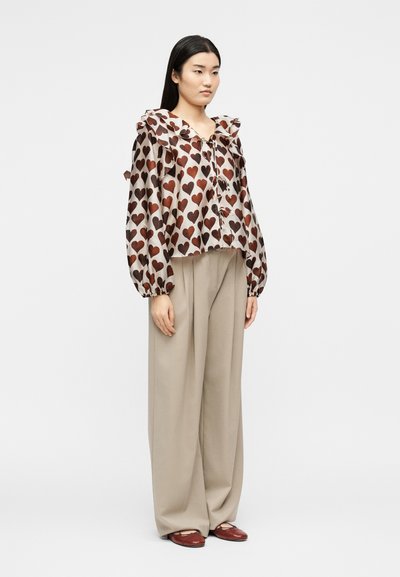 Γυναίκα που στέκεται φορώντας μπεζ παντελόνι με φαρδιά πόδια και μια άνετη μπλούζα με μακριά μανίκια, με μεγάλο βολάν για γιακά και καφέ σχέδια με καρδιές.