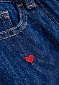 Blåt denimmateriale med orange syninger og et lille broderet rødt hjerte nær lommeområdet.