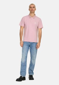 Camisa polo rosa claro con cuello, mangas cortas y un pequeño logo en el pecho. Combinada con jeans de mezclilla azules y zapatos negros.