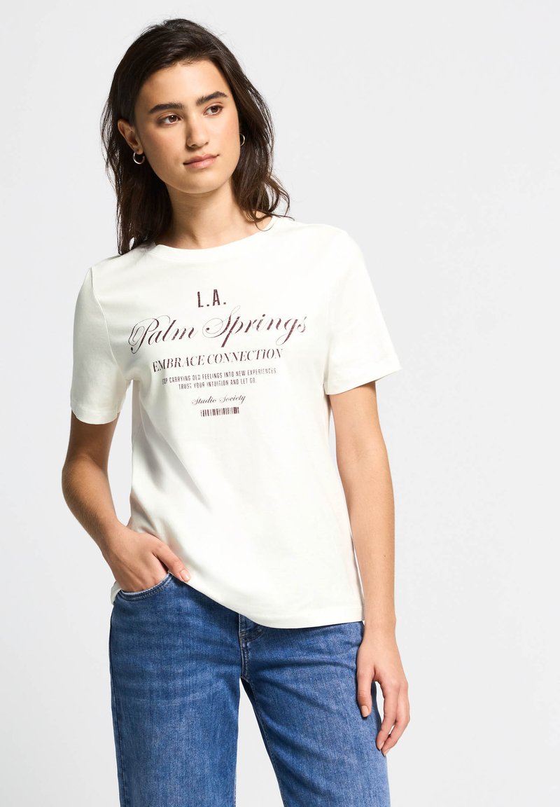 Hvid bomulds-t-shirt med korte ærmer, med et trykt grafisk motiv i brune bogstaver, der står "L.A. Palm Springs Embrace Connection."