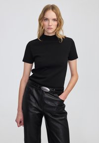 Kobieta z blond włosami ubrana w czarny top z krótkim rękawem i stójką oraz czarne skórzane spodnie z czarnym paskiem i srebrną klamrą.