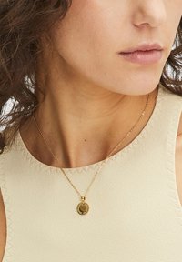 Eine goldfarbene Halskette mit einem runden Anhänger, der ein graviertes Design aufweist, hängt an einer zarten Kette vor einem cremefarbenen Stoffhintergrund.