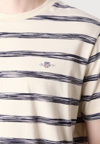 Tricou cu mâneci scurte, în dungi, de culoare crem cu linii bleumarin. Prezintă un logo brodat pe partea stângă a pieptului. Material din bumbac moale, croi lejer.