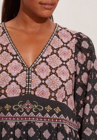 Svart tyg med ett geometriskt mönster av orange, lila och vita former. V-ringad design med broderade detaljer i rosa och grönt.