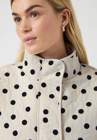 Jeune femme aux cheveux clairs portant une veste matelassée blanche avec de grands pois noirs et des motifs de feuilles texturés.