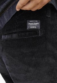 Crne kord pantalone s rebrastom teksturom. Uključuju brendiranu oznaku s natpisom "JACK & JONES" i mali džep na stražnjoj strani.