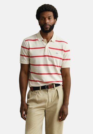 Mann mit Bart, der ein beiges Poloshirt mit roten Streifen und beige Hosen trägt und vor einem schlichten weißen Hintergrund steht.