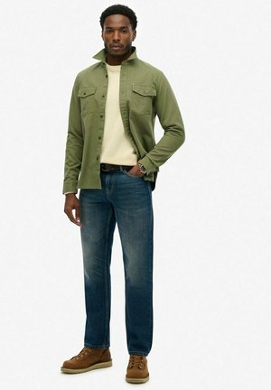 Grüne Button-Down-Hemden über einem cremefarbenen Pullover, blaue Jeans und braune Wanderschuhe. Das Hemd hat zwei Brusttaschen und eine Slim-Fit-Passform.