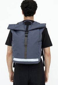 Rulltop ryggsäck i mörkgrått tyg med svarta remmar, reflekterande accent och en minimal logotyp. Strukturerad yta, funktionell design.