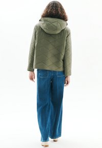 Henkilö, joka on pukeutunut oliivinvihreään päällystakkiin, jossa on huppu, ja leveälahkeisiin sinisiin farkkuihin, seisoo selin valkoista taustaa vasten.