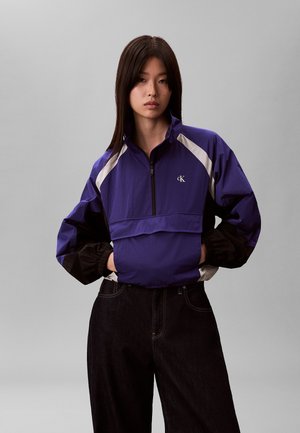 Jonge vrouw met lang donker haar, gekleed in een paarse en zwarte windjack met witte accenten en zwarte broek met hoge taille.