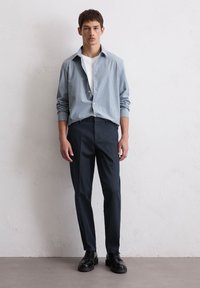 Chemise à rayures bleu clair, t-shirt blanc en dessous, pantalon sombre sur mesure, chaussures noires à lacets, se tenant contre un mur uni.