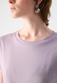 Šviesiai purpurinė marškinėliai su apykakle ir trumpomis rankovėmis. Priekyje yra tekstas, dekoruotas kristalais. Glotni medžiagos tekstūra.