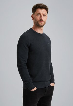 Pull-over noir texturé à col rond, avec des manches longues et des poignets côtelés, porté avec un pantalon sombre. Un logo subtil est visible sur la poitrine.