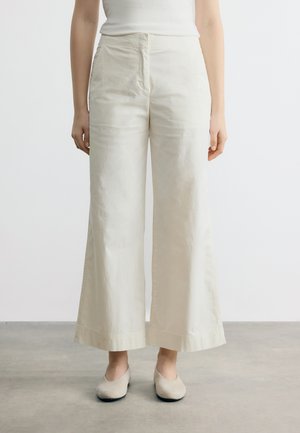 Naine, kes kannab kõrge vöökohaga laia säärega kreemikaid pükse ja beeže ballerinasid, seisab hallil põrandal, valge tühja tausta ees.