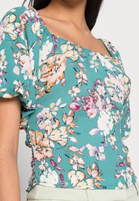 Blusa floreale con scollatura quadrata, maniche a sbuffo corte e tessuto teal morbido con stampa floreale multicolore e dettagli arricciati sui lati.
