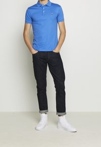 Mies, joka on pukeutunut siniseen lyhythihaiseen polo-paitaan, tummiin käärityisiin farkkuihin ja valkoisiin korkeatennareihin, seisoo kädet taskuissa.