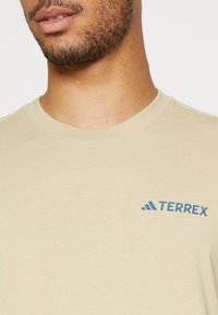 Beige bomulds-t-shirt med rund halsudskæring, korte ærmer og et blåt og turkis logo på venstre bryst. Glat tekstur.