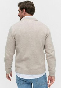 Beige stickad tröja med sjalkrage, ribbade ärmslut och nederkant. Bakvy visar strukturerad tyg, avslappnad passform och enkel design.