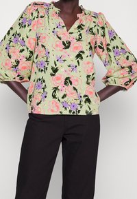 Blouse fleurie vert clair avec motifs floraux roses et violets, col froncé et manches bouffantes. Portée avec un pantalon noir.