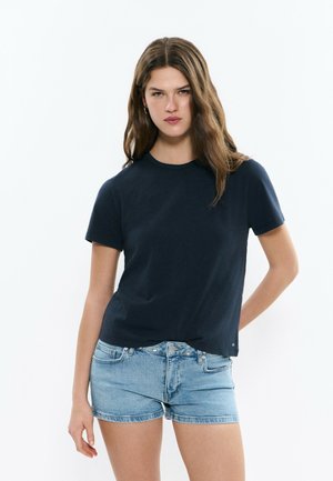 Jeune femme aux longs cheveux bruns portant un T-shirt à manches courtes bleu marine foncé et un short en jean bleu clair sur un fond blanc uni.