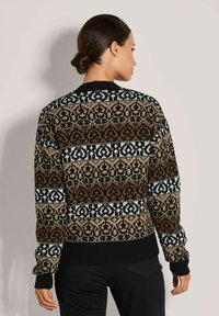 Frau mit dunklem Haar in einem tiefen Dutt, die einen schwarz-braun-weißen gemusterten Strickpullover und schwarze Hosen trägt, steht vor einer schlichten Wand.