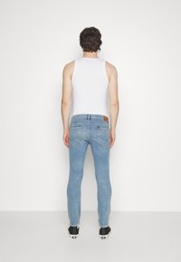 Jeans ajustados de color azul claro con un parche de cuero de la marca en la parte trasera, llevados con una camiseta sin mangas blanca y zapatos negros, mostrados desde la parte trasera.
