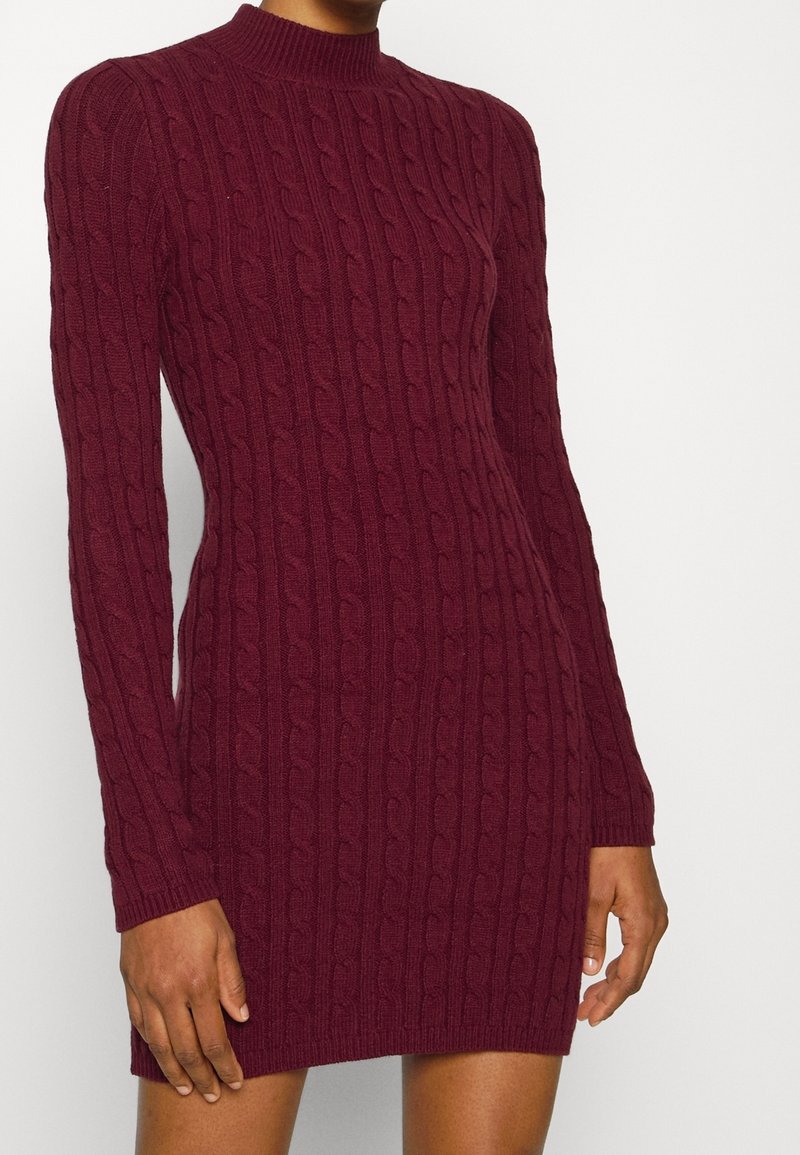 Gros plan d'une personne portant une robe-pull bordeaux ajustée à manches longues avec col montant en maille torsadée.