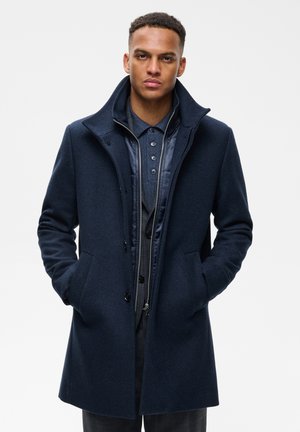 Homme portant un manteau bleu marine superposé, une veste et une chemise à col, les mains dans les poches du manteau, sur un fond clair uni.
