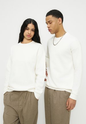 Deux jeunes adultes portant des hauts blancs à manches longues et des pantalons à carreaux marron se tiennent côte à côte devant un fond uni.