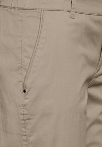 Pantaloni di cotone beige con una texture liscia, dotati di una tasca laterale e un piccolo dettaglio in bottone nero vicino all'apertura della tasca.
