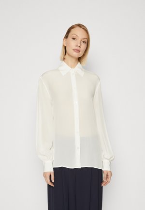 Camicia bianca trasparente con colletto, maniche lunghe a sbuffo e polsini abbottonati. Il tessuto sembra leggero e con una superficie liscia.