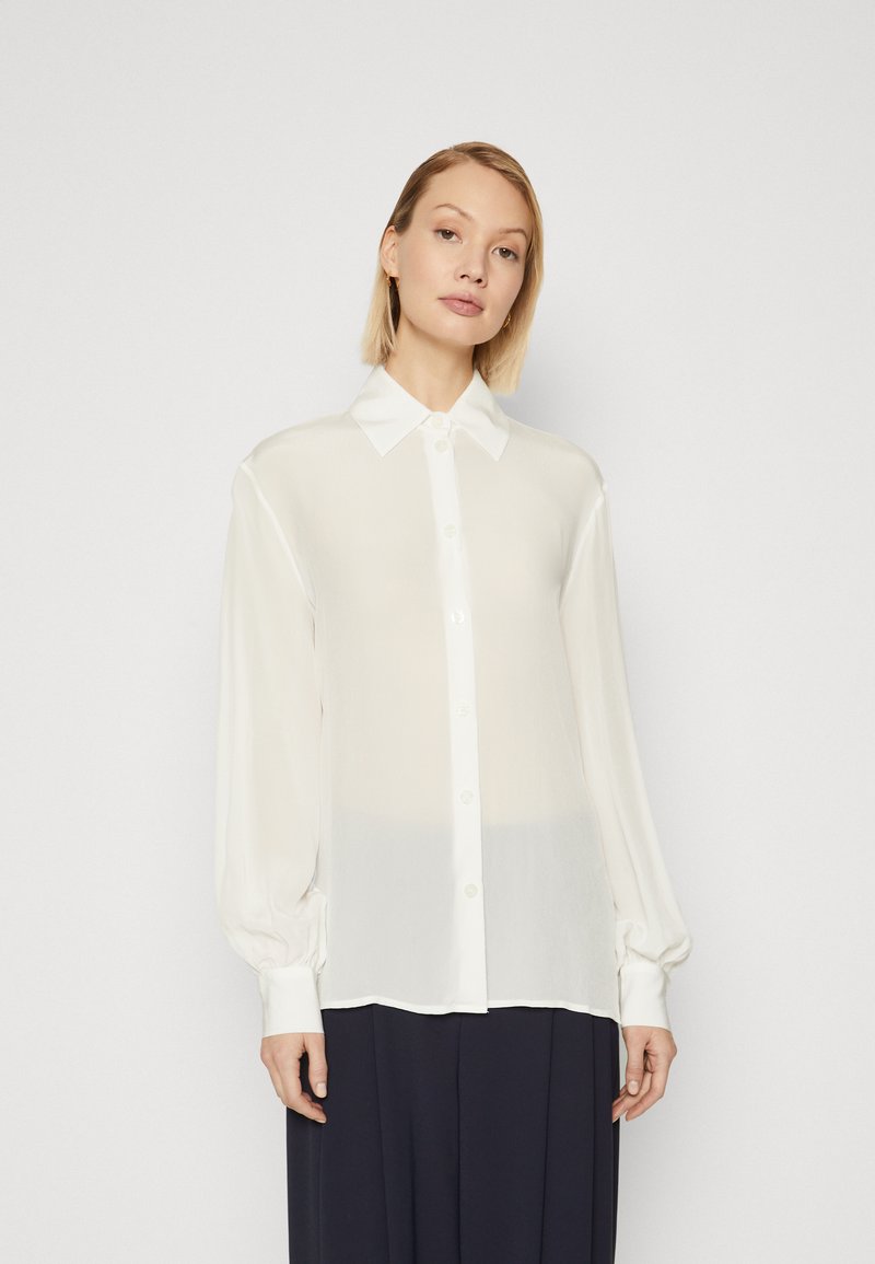 Camicia bianca trasparente con colletto, maniche lunghe a sbuffo e polsini abbottonati. Il tessuto sembra leggero e con una superficie liscia.