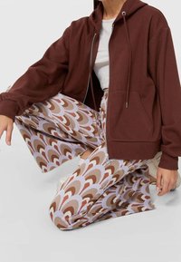 Bruine zip-up hoodie met grote zakken, gecombineerd met brede broek met een patroon in roze, bruin en crèmekleurige golfontwerp, gedragen met witte sneakers.