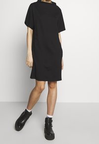 Svart oversized klänning med hög hals, korta ärmar och en avslappnad passform, tillverkad av mjukt tyg, tillsammans med kraftiga svarta ankelboots.