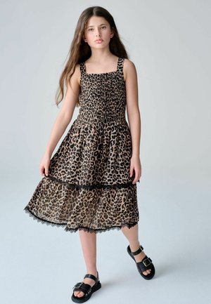 Meisje met lang bruin haar, gekleed in een mouwloze jurk met luipaardprint en zwarte sandalen met gespen, staand tegen een lichtgrijze achtergrond.