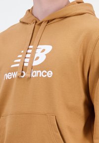 Sudadera con capucha marrón, hecha de tela suave, con un bolsillo tipo canguro en la parte frontal y un logo blanco de New Balance en el pecho.
