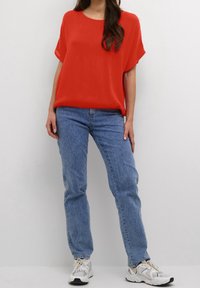 Rotes kurzärmliges Oberteil mit Rundhalsausschnitt, kombiniert mit hellblauen geraden Jeans und weißen Sneakers. Schlichte Styling, keine sichtbaren Muster.