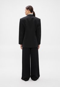 Μαύρο μεγάλο σακάκι με γιακά με σχισμή, συνδυασμένο με παντελόνια φαρδιάς γραμμής, λείο ύφασμα, μινιμαλιστικός σχεδιασμός, χωρίς ορατά σχέδια ή διακοσμήσεις.
