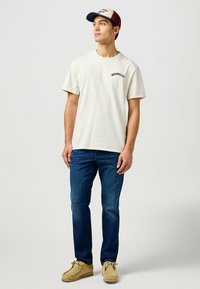 Cremefarvet t-shirt med korte ærmer og "WRANGLER" logo, parret med blå jeans og beige mokkasiner, toppet med en tofarvet kasket.