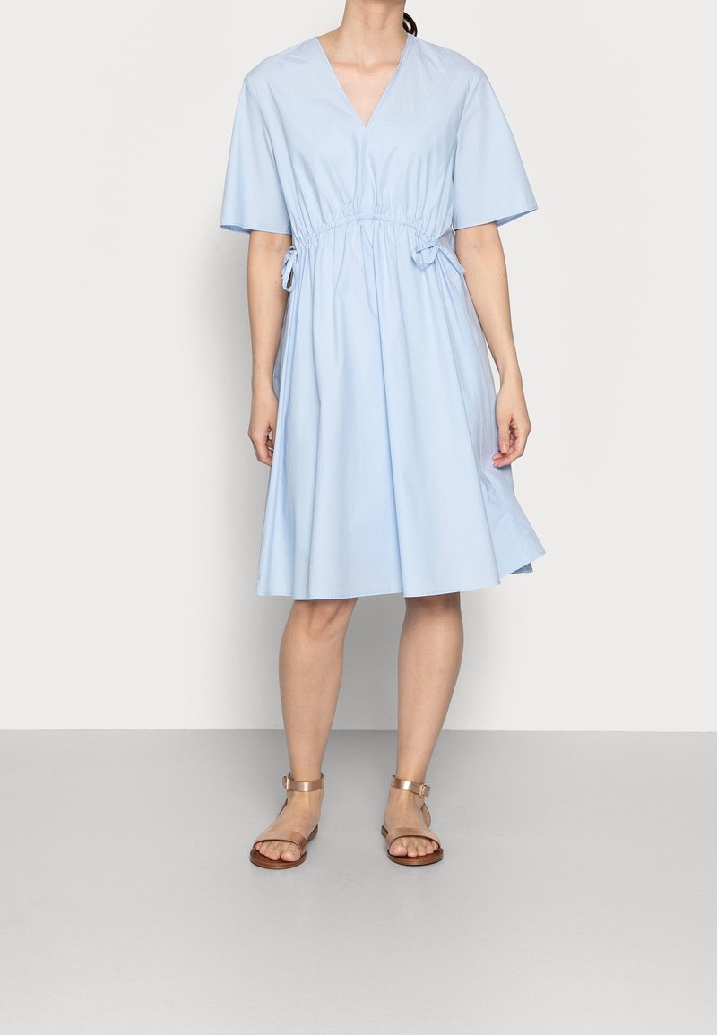Femme portant une robe bleu clair longueur genou avec manches courtes et liens latéraux, associée à des sandales plates métalliques à bride cheville.