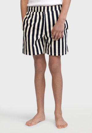 Personne portant un short à rayures verticales noires et blanches avec une ceinture élastique, debout pieds nus sur un fond uni.