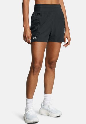 Kvinde iført sorte Under Armour træningsshorts, hvide sokker og hvide sneakers, stående mod en ensfarvet baggrund.