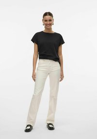 Camiseta negra de manga corta combinada con unos pantalones vaqueros de campana beige claro y mocasines negros. El modelo está de pie frente a un fondo blanco liso.