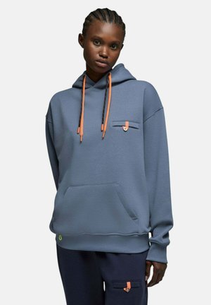 Personne portant un sweat à capuche bleu avec des cordons orange et un pantalon assorti avec des poches à boutons orange, debout devant un fond uni.