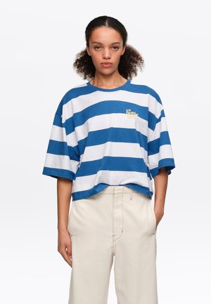 Vrouw met krullend haar die een losse blauw-wit gestreepte korte mouwen shirt en lichtbeige broek draagt, met handen in de zakken.