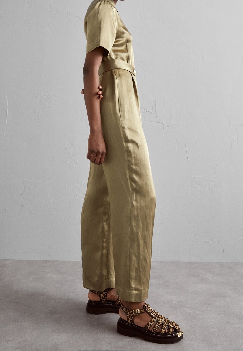 Salopette vert olive à manches courtes avec taille ceinturée et poches latérales, assortie à des sandales brunes à motif léopard dotées d'un design de lanière.