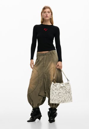 Žena koja nosi crnu bluzu s dugim rukavima i crvenim detaljem, široke maslinasto zelene hlače, crne čizme, drži bijelu tkanu torbu s vezicom.