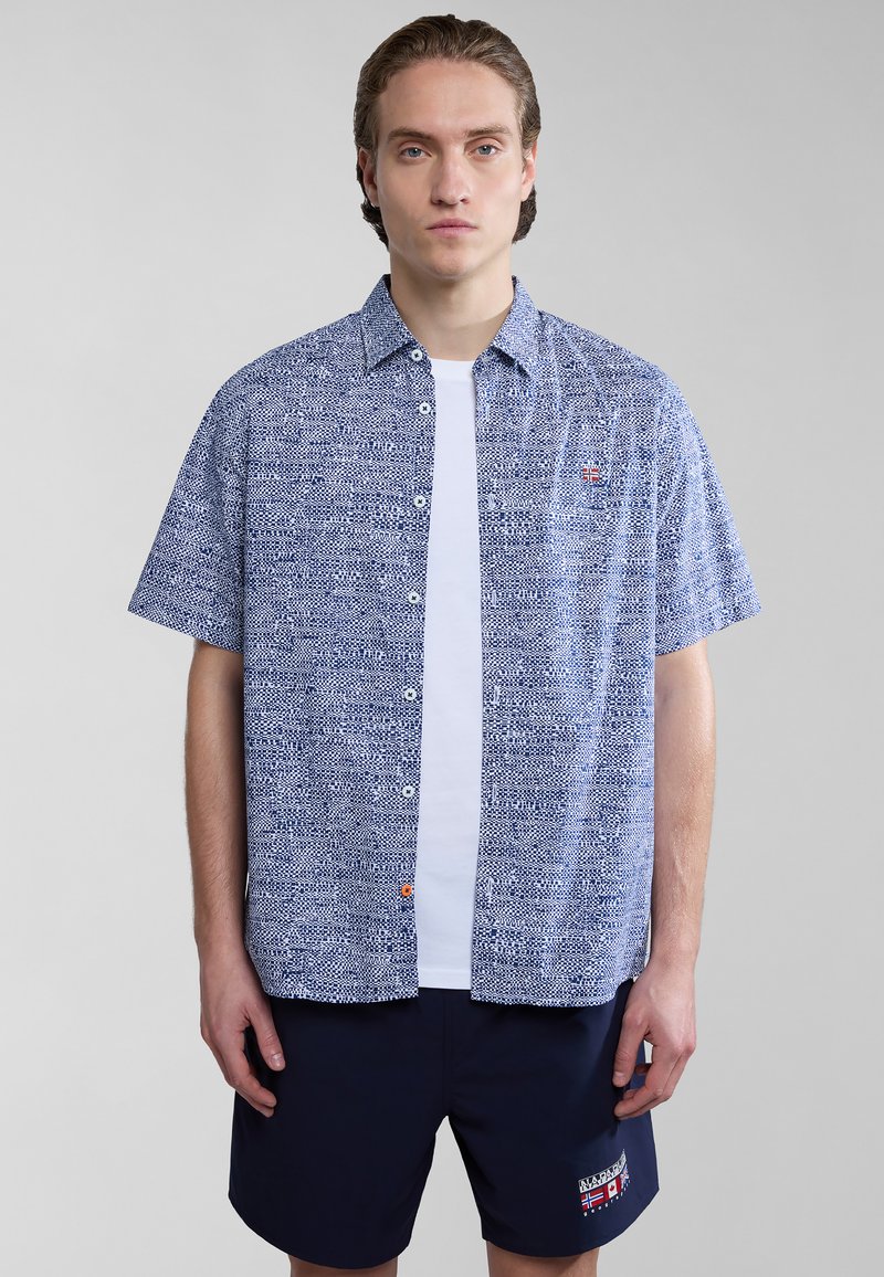 Camisa de botones de manga corta en azul marino y blanco con un patrón geométrico; cuenta con un logo en el pecho y se combina con pantalones cortos azul marino.